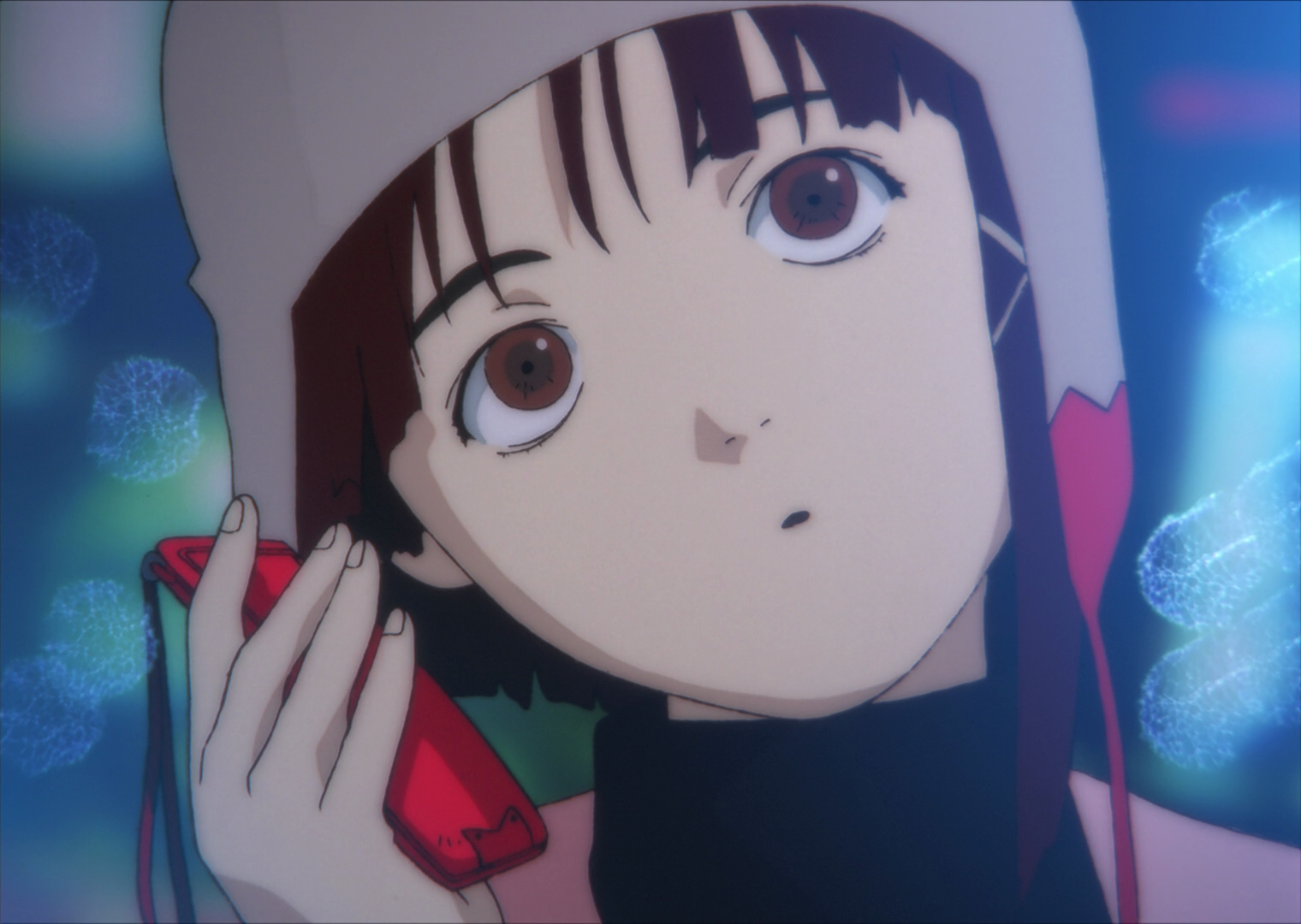 Lain Iwakura stands in the center of frame, her head tilted to the left. She has brown eyes, reddish-brown hair which has a long bang near the right side of her face which is tied in a thick, dense strand, by an X-shaped clip. She is wearing a black turtleneck sweater with a pink jacket over it, also wearing a gray winter hat with red ear flaps. She is holding a red mobile phone with a matching red wrist strap lanyard attached to it. She is surrounded by bright lights as she walks down the street.